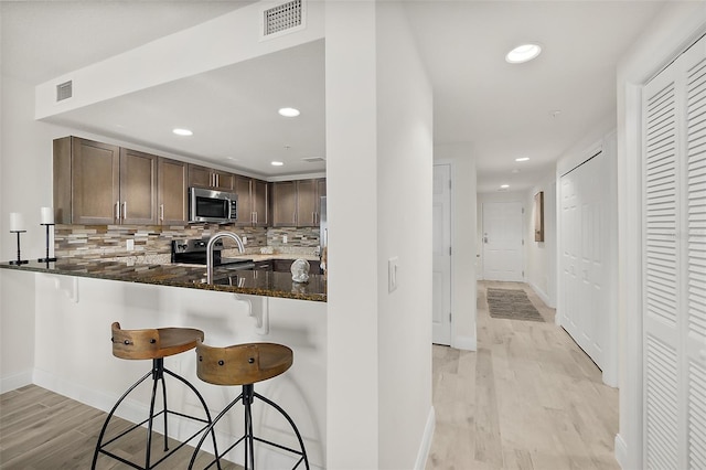 kitchen with kitchen peninsula, a kitchen bar, dark stone counters, stainless steel appliances, and light hardwood / wood-style floors