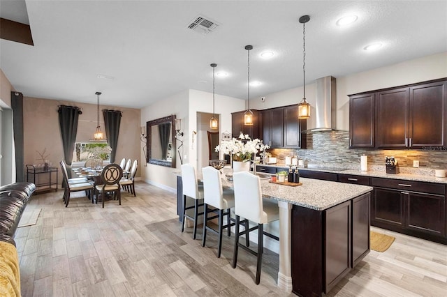 kitchen with wall chimney exhaust hood, hanging light fixtures, a kitchen island with sink, a breakfast bar, and dark brown cabinets