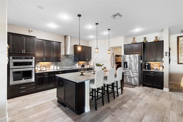 kitchen with pendant lighting, a kitchen island with sink, a breakfast bar area, wall chimney exhaust hood, and stainless steel appliances