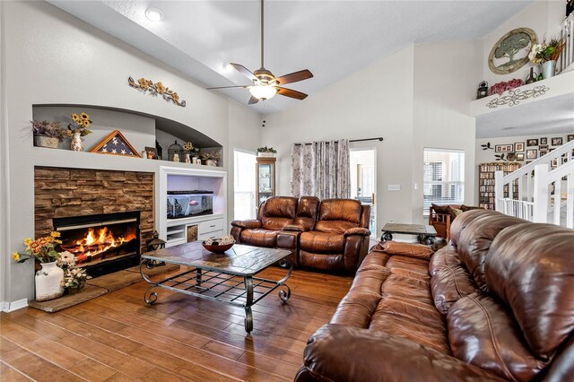 living room with built in shelves, ceiling fan, a stone fireplace, wood-type flooring, and high vaulted ceiling