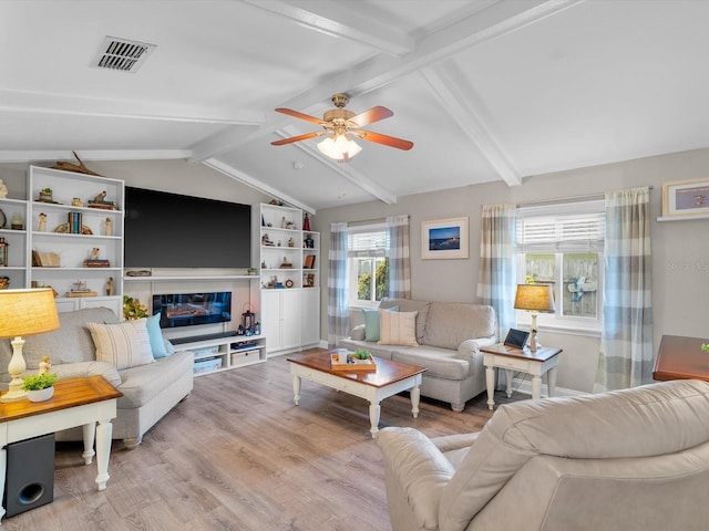 living room featuring lofted ceiling with beams, ceiling fan, wood finished floors, visible vents, and a glass covered fireplace