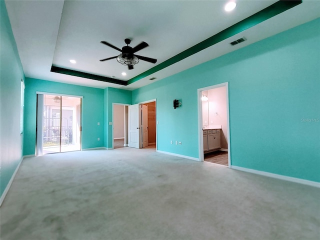 unfurnished bedroom featuring ceiling fan, ensuite bathroom, a tray ceiling, and light carpet