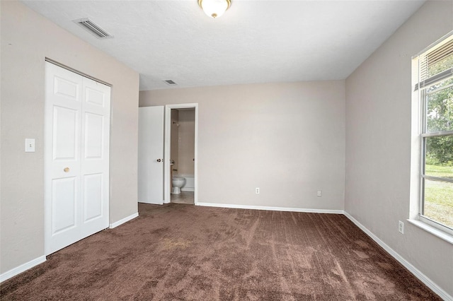 unfurnished bedroom featuring dark carpet, a closet, ensuite bathroom, and multiple windows