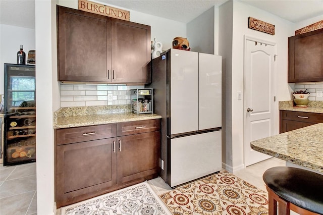 kitchen with light stone counters, backsplash, freestanding refrigerator, light tile patterned flooring, and dark brown cabinets