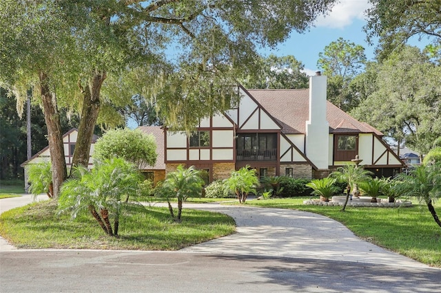 tudor-style house with brick siding, driveway, stucco siding, a front lawn, and a chimney