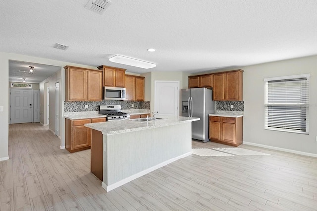 kitchen featuring an island with sink, light wood-type flooring, decorative backsplash, stainless steel appliances, and a textured ceiling