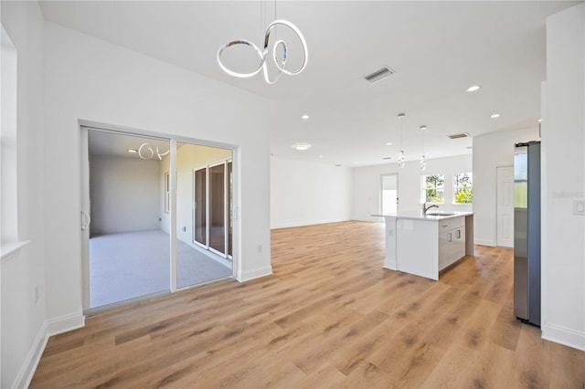 interior space featuring stainless steel refrigerator, sink, an island with sink, pendant lighting, and light hardwood / wood-style floors