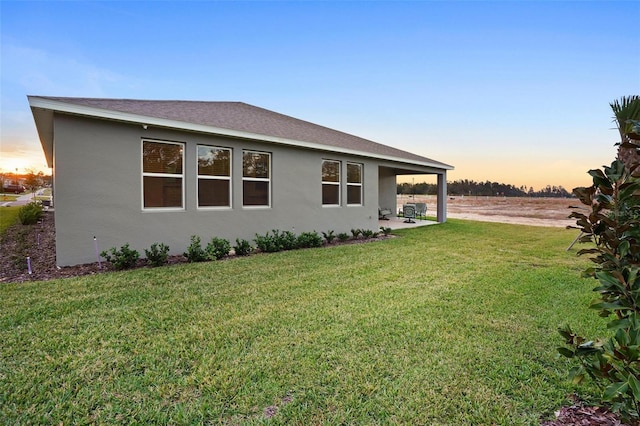 property exterior at dusk with a shingled roof, a patio area, a yard, and stucco siding