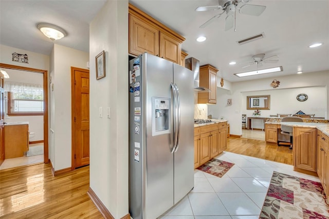 kitchen featuring light stone countertops, ceiling fan, wall chimney exhaust hood, stainless steel appliances, and light hardwood / wood-style floors