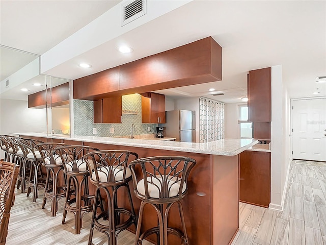 kitchen with kitchen peninsula, stainless steel fridge, decorative backsplash, a kitchen breakfast bar, and light hardwood / wood-style flooring