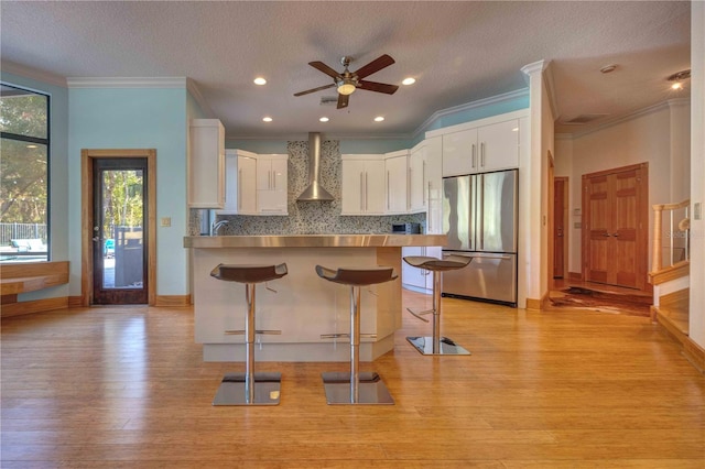 kitchen with wall chimney exhaust hood, stainless steel built in refrigerator, light hardwood / wood-style flooring, white cabinetry, and a breakfast bar area