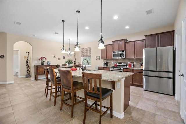 kitchen featuring stainless steel appliances, light stone counters, an island with sink, decorative light fixtures, and a kitchen bar