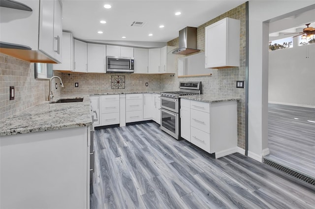 kitchen featuring wall chimney range hood, sink, ceiling fan, appliances with stainless steel finishes, and white cabinetry