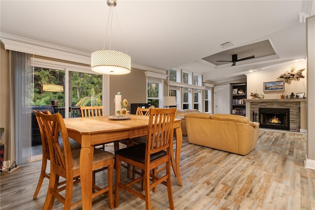 dining area featuring a lit fireplace, a ceiling fan, light wood finished floors, a raised ceiling, and ornamental molding