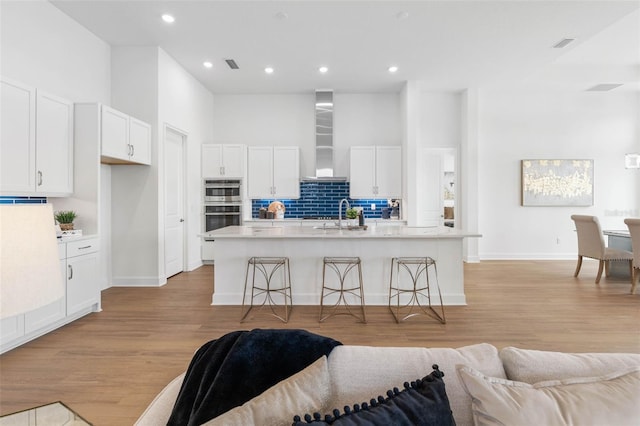 kitchen featuring wall chimney exhaust hood, white cabinets, light hardwood / wood-style flooring, and an island with sink