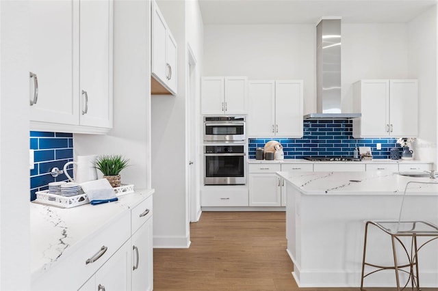 kitchen featuring white cabinets, wall chimney exhaust hood, double oven, light hardwood / wood-style floors, and light stone counters