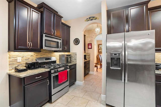 kitchen featuring dark brown cabinetry, light stone countertops, stainless steel appliances, backsplash, and light tile patterned floors