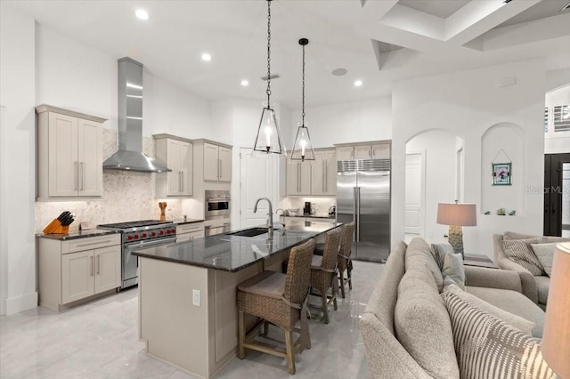 kitchen with wall chimney range hood, sink, decorative backsplash, an island with sink, and premium appliances