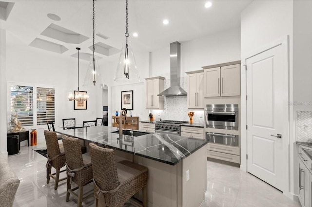 kitchen with a towering ceiling, an island with sink, stainless steel appliances, and wall chimney range hood