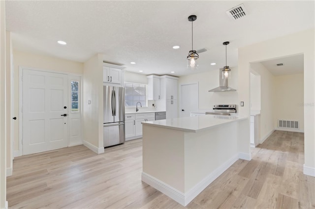 kitchen featuring white cabinets, wall chimney range hood, light hardwood / wood-style flooring, decorative light fixtures, and stainless steel appliances