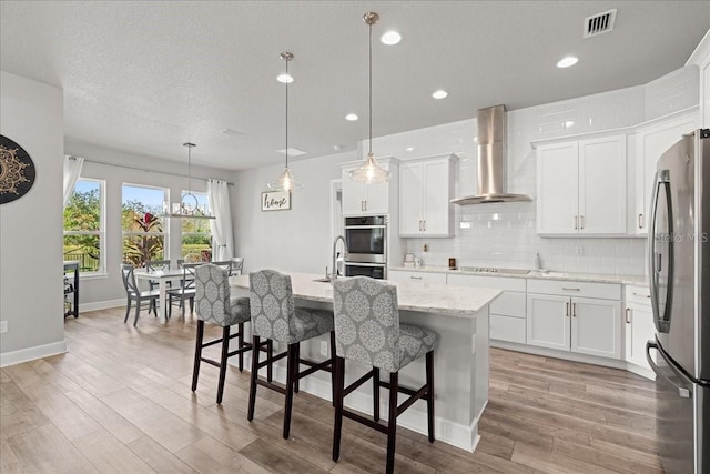 kitchen featuring wall chimney exhaust hood, hanging light fixtures, a kitchen island with sink, white cabinets, and appliances with stainless steel finishes