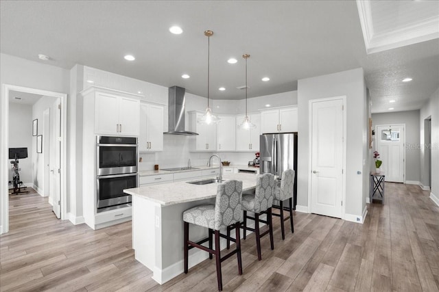 kitchen featuring white cabinetry, wall chimney exhaust hood, hanging light fixtures, a center island with sink, and appliances with stainless steel finishes