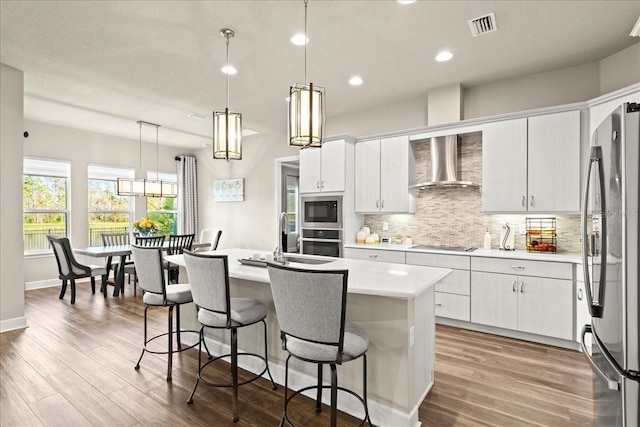 kitchen featuring white cabinetry, stainless steel appliances, wall chimney range hood, an island with sink, and pendant lighting