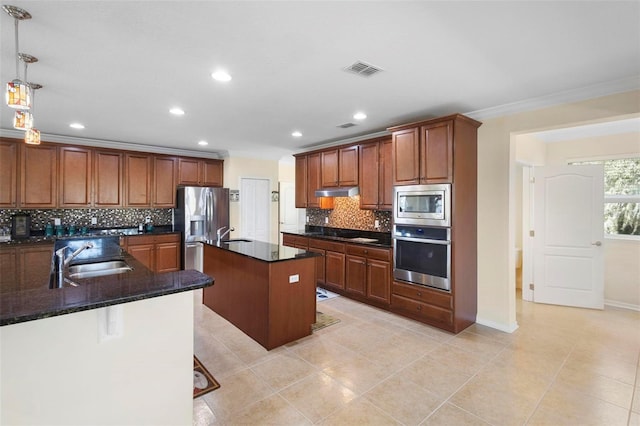 kitchen with sink, appliances with stainless steel finishes, a kitchen island with sink, hanging light fixtures, and dark stone countertops