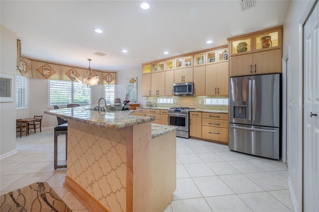 kitchen with pendant lighting, a kitchen island with sink, stainless steel appliances, light brown cabinetry, and light stone counters