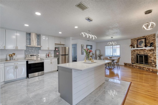 kitchen featuring a textured ceiling, stainless steel appliances, wall chimney range hood, white cabinets, and hanging light fixtures