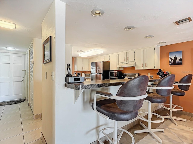 kitchen with light tile patterned floors, kitchen peninsula, stainless steel fridge, a breakfast bar area, and white cabinets