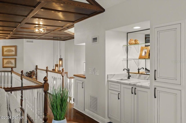 bar with light stone counters, coffered ceiling, sink, decorative light fixtures, and white cabinetry