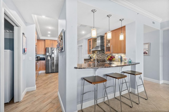 kitchen featuring hanging light fixtures, wall chimney range hood, stainless steel refrigerator with ice dispenser, kitchen peninsula, and dark stone counters