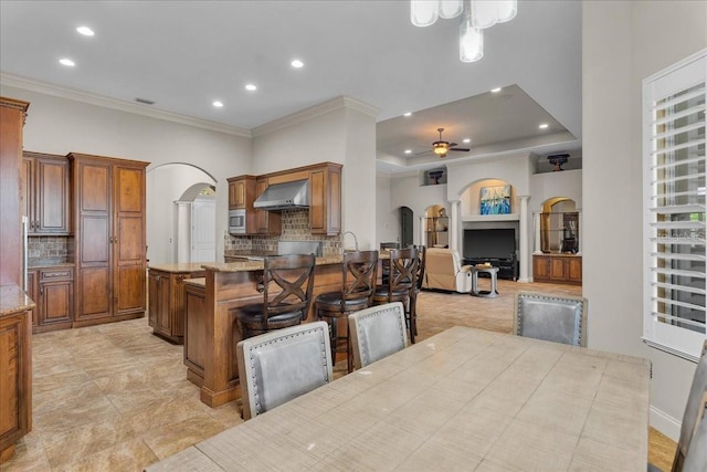 kitchen featuring arched walkways, brown cabinets, stainless steel microwave, and wall chimney range hood