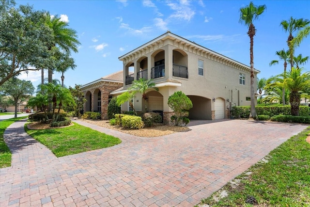 italianate-style house with an attached garage, a balcony, decorative driveway, and stucco siding