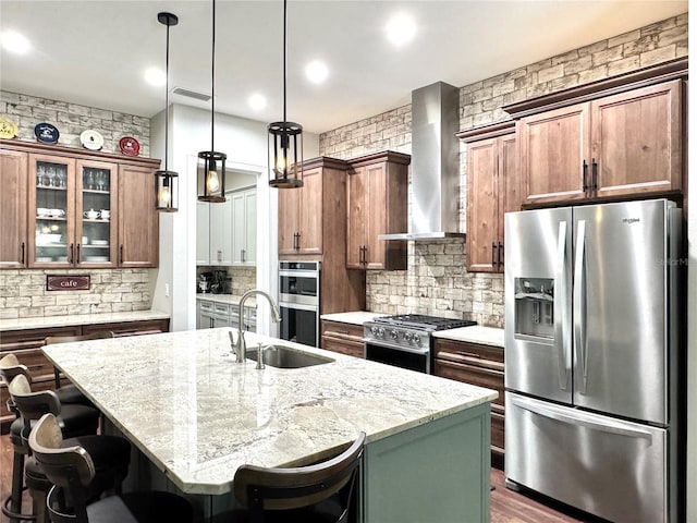 kitchen with appliances with stainless steel finishes, an island with sink, light stone countertops, wall chimney range hood, and decorative light fixtures