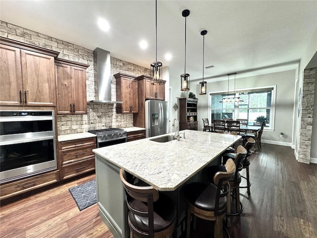 kitchen featuring wall chimney exhaust hood, hanging light fixtures, an island with sink, appliances with stainless steel finishes, and a kitchen breakfast bar