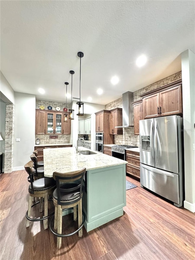 kitchen featuring sink, light stone counters, wall chimney range hood, a kitchen island with sink, and appliances with stainless steel finishes