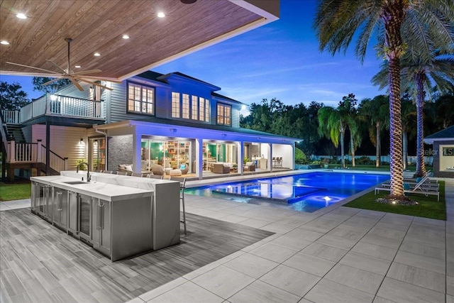 pool at dusk featuring ceiling fan, an outdoor wet bar, and exterior kitchen