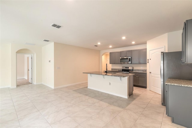 kitchen featuring appliances with stainless steel finishes, a kitchen bar, stone countertops, gray cabinets, and a center island with sink