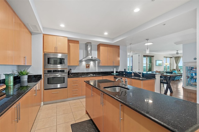 kitchen with a center island with sink, sink, appliances with stainless steel finishes, wall chimney exhaust hood, and dark stone counters