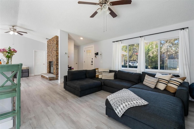 living room with ceiling fan, a textured ceiling, a stone fireplace, vaulted ceiling, and light wood-type flooring