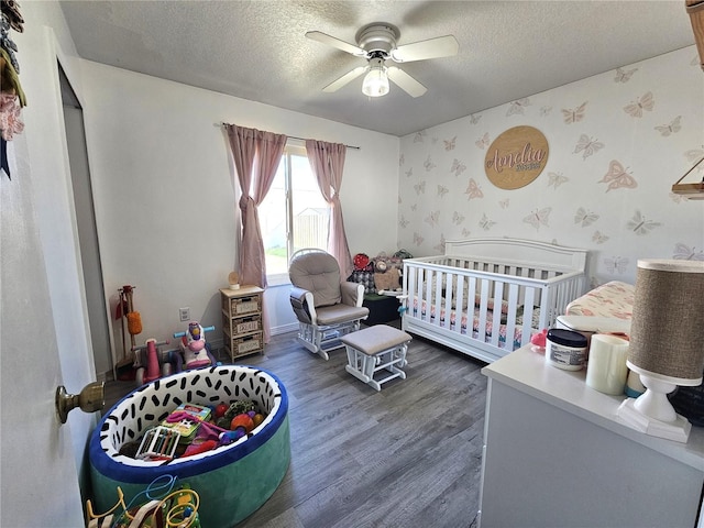 bedroom with a textured ceiling, dark wood-type flooring, a nursery area, and ceiling fan