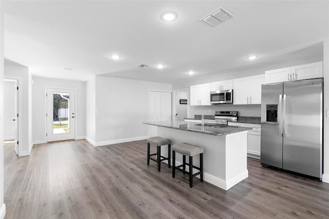 kitchen featuring white cabinetry, stainless steel appliances, an island with sink, dark hardwood / wood-style flooring, and dark stone counters