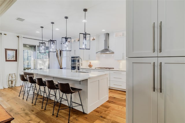 kitchen featuring pendant lighting, white cabinetry, wall chimney range hood, light stone counters, and a center island with sink