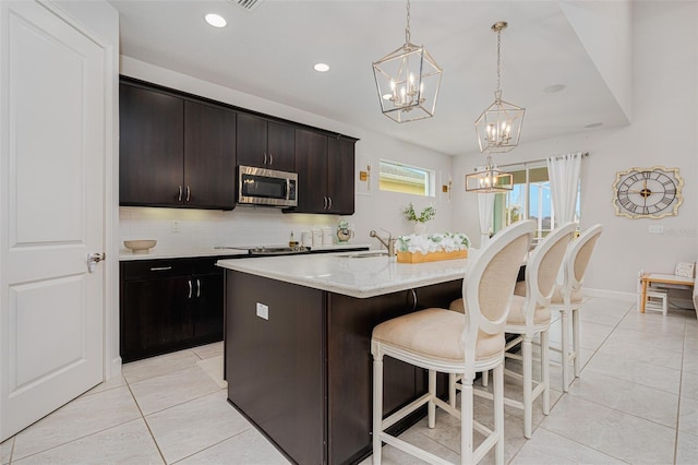 kitchen featuring dark brown cabinetry, tasteful backsplash, hanging light fixtures, light tile patterned floors, and an island with sink