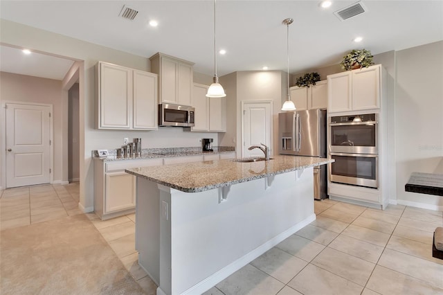 kitchen with sink, appliances with stainless steel finishes, a kitchen island with sink, hanging light fixtures, and light stone counters