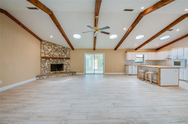 unfurnished living room featuring sink, light hardwood / wood-style flooring, ceiling fan, lofted ceiling with beams, and a stone fireplace