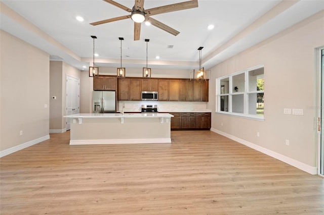 kitchen with appliances with stainless steel finishes, hanging light fixtures, light hardwood / wood-style floors, a raised ceiling, and a center island with sink