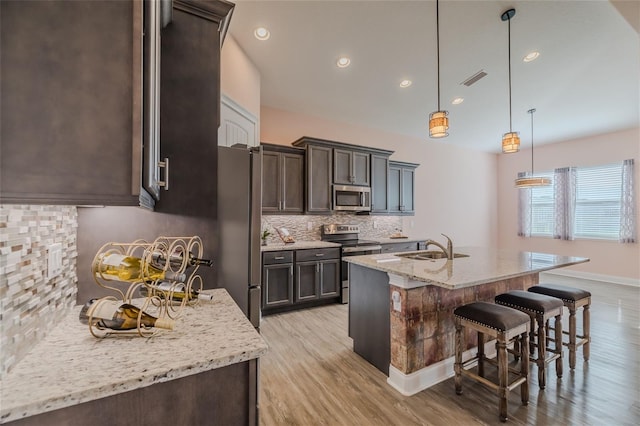 kitchen with a kitchen island with sink, stainless steel appliances, light stone countertops, dark brown cabinets, and pendant lighting